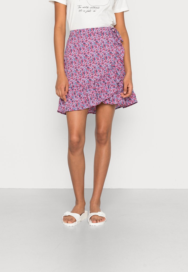 Femme portant une jupe portefeuille fleurie rose et violette avec ourlet à volants, un t-shirt blanc à motif graphique et des sandales blanches à enfiler, debout devant un fond uni.