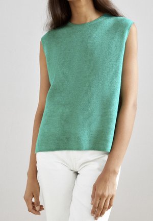 Vrouw draagt een mouwloze, mintgroene gebreide top en witte broek, staande tegen een effen lichte achtergrond.