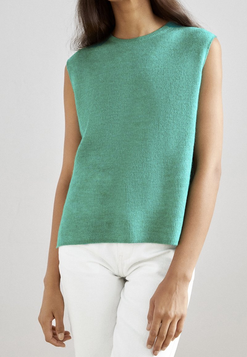 Femme portant un haut en tricot sans manches vert menthe et un pantalon blanc, debout devant un fond clair uni.