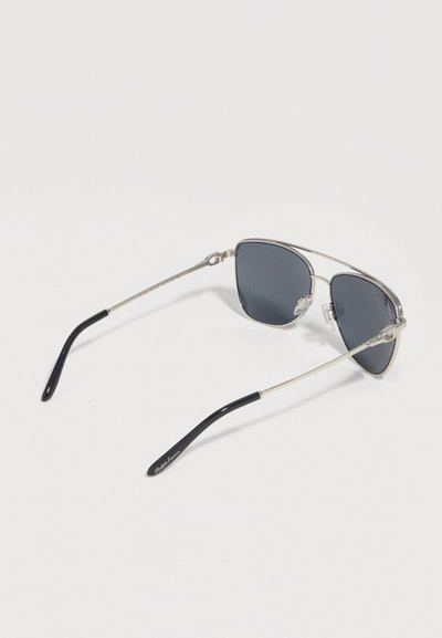 Γυαλιά ηλίου με ασημένιο μεταλλικό σκελετό, σκούρους φακούς και μαύρα πλαστικά άκρα στους βραχίονες, ακουμπισμένα με τους φακούς προς τα κάτω πάνω σε λευκή επιφάνεια.