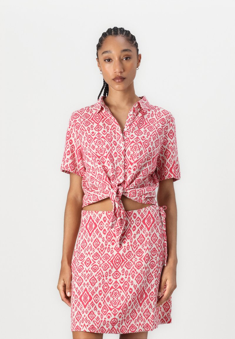 Completo gonna e blusa a maniche corte con nodo frontale in rosa e bianco, caratterizzato da motivi geometrici e un tessuto morbido e texturizzato.