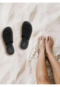 Sandales noires à enfiler avec un design texturé à rainures ; dotées d'une semelle intérieure rembourrée. Sable sous les pieds dans un cadre de plage clair.