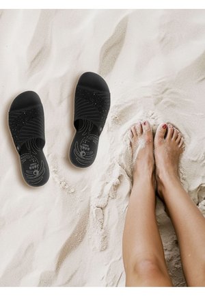 Sandales noires à enfiler avec un design texturé à rainures ; dotées d'une semelle intérieure rembourrée. Sable sous les pieds dans un cadre de plage clair.