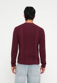 Suéter tricotado em burdeos com padrões de trança e diamante, gola redonda e punhos e bainha canelados. Usado com jeans de denim azul claro.