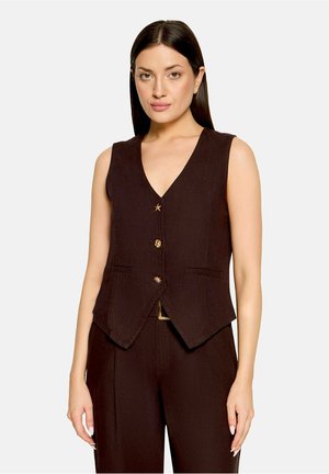 Femme portant un gilet marron foncé sans manches à col en V avec des boutons décoratifs et un pantalon assorti, debout devant un fond clair uni.