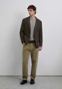 Un joven vestido con una chaqueta de cuadros marrón, un suéter beige, pantalones caqui y zapatos negros, de pie contra una pared gris claro y lisa.