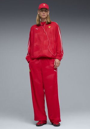 Persona in piedi che indossa una tuta da corsa rossa Puma Ferrari, cappellino abbinato e auricolari, su uno sfondo grigio uniforme.