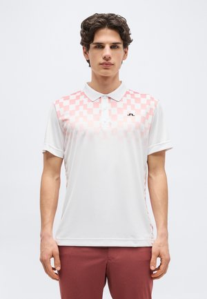 Νεαρός άνδρας με κοντά σγουρά μαλλιά, που φοράει λευκό πόλο πουκάμισο με ροζ καρό μοτίβο στο πάνω μέρος του στήθους και μπορντό παντελόνι.