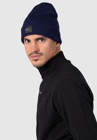 Beanie lavorato a maglia blu navy con texture a coste, risvolto pieghevole e una piccola etichetta nera, indossato da un modello in un dolcevita nero.