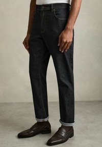Mann trägt dunkle hochgekrempelte Jeans mit braunen Loafers und dunklen Socken, steht auf einem hellgrauen Boden vor einer beigen Wand.