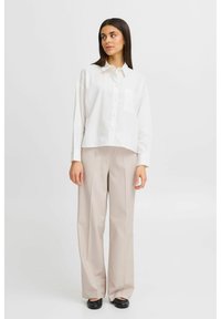 Camicia bianca abbottonata corta con taschino sul petto, abbinata a pantaloni larghi beige chiaro e scarpe nere slip-on. Design semplice.