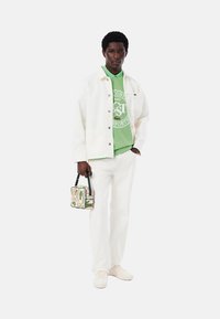 Chaqueta de mezclilla blanca, camiseta gráfica verde, pantalones blancos y zapatillas ligeras; sosteniendo una bolsa estampada con acentos verdes y rojos.