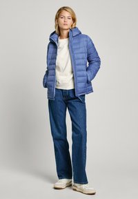 Lichtblauwe gewatteerde jas met capuchon, ritssluiting en een doorgestikt patroon, gecombineerd met een crèmekleurige top en wijde blauwe jeans. Witte sneakers.