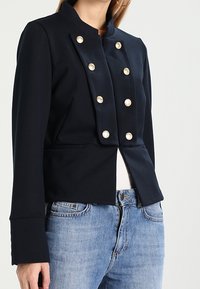 Marinblå strukturerad jacka med hög krage, försedda med sex stora pärlliknande knappar framtill och en kortad design.