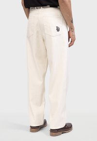 Un pantalon blanc à jambes droites avec des poches arrière et une patch logo. Fabriqué en tissu durable avec des coutures visibles et une coupe décontractée.