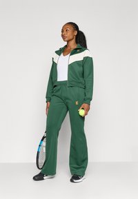 Tuta verde realizzata in tessuto morbido; giacca con zip e dettagli crema, pantaloni a vita alta e logo sul dettaglio. Tiene in mano una palla da tennis e una racchetta.