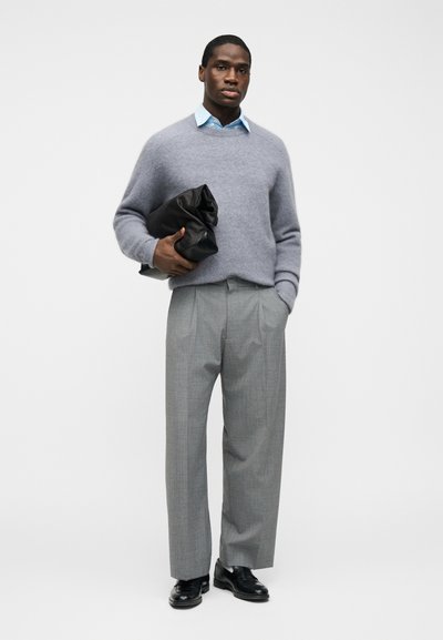 Άνδρας με γαλάζιο πουκάμισο, γκρι πουλόβερ, γκρι πλισέ παντελόνι και μαύρα παπούτσια κρατώντας μια μεγάλη μαύρη διπλωμένη τσάντα φάκελο σε λευκό φόντο.