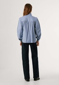 Modrá a biela pruhovaná blúzka s balónovými rukávmi a mandarínským golierom, kombinovaná s tmavými džínsami. Detail predného skladania.