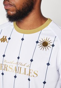Valkoinen urheilupaita, jossa on kultaisia yksityiskohtia, timanttikuvio, Kappa-logo ja "Football Club de Versailles" -teksti, pyöreä kaulus.