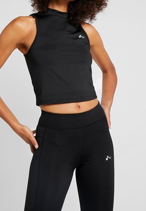 Femme portant un crop top noir ajusté sans manches et des leggings noirs taille haute avec petits logos blancs sur chaque vêtement.