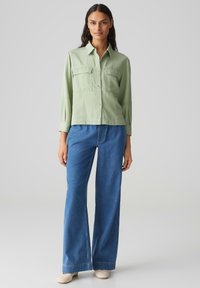 Hellgrünes Hemd mit Knopfverschluss und zwei Fronttaschen, kombiniert mit weiten blauen Jeans. Das Hemd hat lange Ärmel und einen lässigen Schnitt.