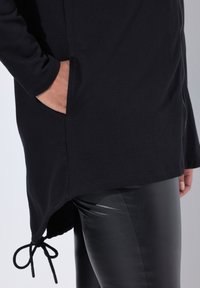 Túnica negra con mangas largas, que cuenta con un bolsillo, un dobladillo curvado y un detalle de cordón en la parte inferior. Combinada con leggings negros brillantes.