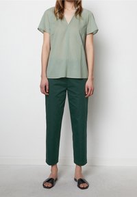 Donna che indossa un top a maniche corte verde chiaro con scollo a V, pantaloni dritti verdi scuri e sandali neri aperti sulla punta, in piedi contro un muro bianco.