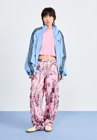 Chaqueta de chándal azul claro con cremallera y tres rayas negras, combinada con pantalones anchos de pierna amplia de patrón rosa. El modelo lleva zapatillas de deporte beige.