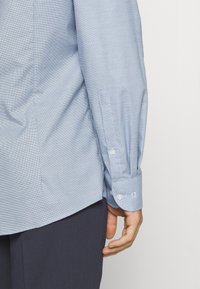 Primo piano di una persona che indossa una camicia a maniche lunghe azzurro chiaro con motivo, con i polsini abbottonati e pantaloni scuri, su uno sfondo neutro.