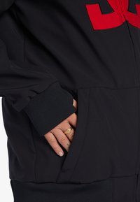 Čierny športový overal s červeným číslom, rebrovanými manžetami a bočnou vreckom s rukou odloženou vo vnútri. Hladká textúra látky.