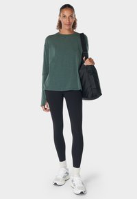 Vrouw in een groene lange mouw top, zwarte leggings, witte sneakers en sokken, met een zwarte schoudertas op een witte achtergrond.