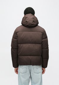 Καφέ μπουφάν puffer με κουκούλα, επενδυμένο σχέδιο και ριμπ μανσέτες, συνδυασμένο με ανοιχτό μπλε τζιν. Φαίνεται ζεστό και ανθεκτικό.