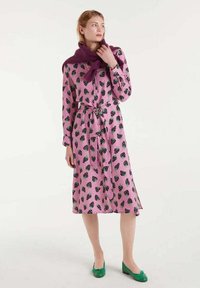 Vestido rosa con patrón de fresas negras, mangas largas, botones en la parte delantera y cinta en la cintura. Combinado con zapatos planos verdes y una bufanda morada.