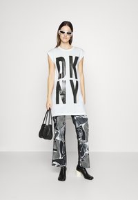 Balta per didelė marškinė su juodu „DKNY“ užrašu, derinama su plačiomis juodai-pilkos raštuotos kelnėmis. Modelis laiko juodą rankinę, vilki baltus akinius nuo saulės.