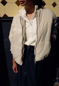 Personne portant une veste matelassée crème par-dessus un pull blanc boutonné et une jupe texturée noire, debout devant un mur carrelé et sombre.