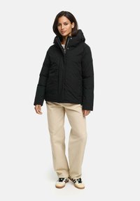 Black padded jacket with a high collar and hood, featuring a zipper closure and side pockets, paired with light beige trousers and shoes.