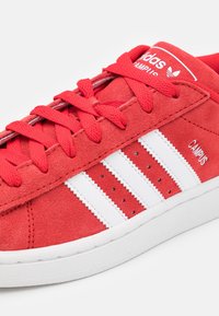 Červené tenisky Adidas Campus s bílými pruhy, červenými tkaničkami a b ílou podrážkou, zachycující bok, jazyk a detaily značky.