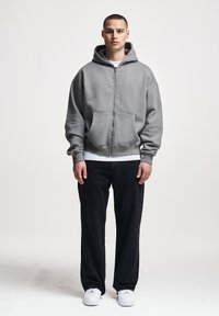 Joven de pie, vistiendo una sudadera con capucha gris con cremallera, camiseta blanca, pantalones negros y zapatillas blancas contra un fondo claro y liso.