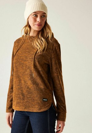 Mujer vestida con una sudadera con capucha marrón texturizada con cordones, jeans azul oscuro y un gorro de punto blanco, de pie frente a un fondo liso y claro.