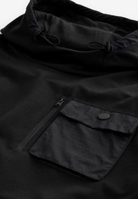 Veste noire avec une poche poitrine zippée et une poche à rabat avec bouton-pression, dotée d'une capuche ajustable avec cordons de serrage.