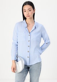 Light blue button-up shirt with vertical stripes, two chest pockets, and gold buttons, paired with a silver, crescent-shaped bag.