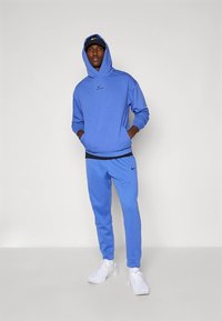 Sweat à capuche bleu avec poche avant et logo noir, assorti à un jogging bleu coordonné. Stylisé avec des baskets blanches et une casquette noire.
