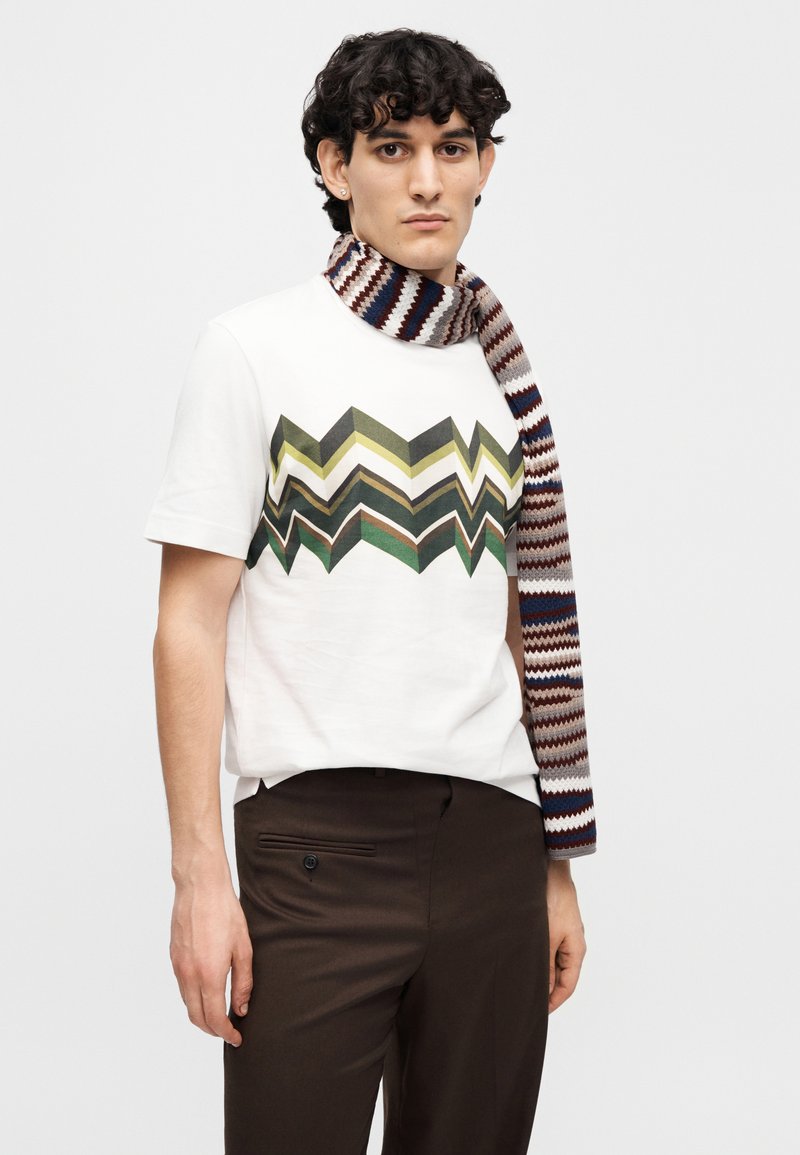 White t-shirt with a zigzag pattern in green, gray, and brown; paired with a multicolored striped scarf. Brown pants with a side pocket.