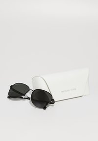Lunettes de soleil noires avec des verres surdimensionnés et angulaires, montées sur une fine monture. Accompagnées d'une écrin blanc avec le logo en relief "MICHAEL KORS".