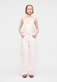 Γυναίκα με κοντά κόκκινα μαλλιά στέκεται με τα χέρια στις τσέπες, φορά διάφανο λευκό καμισόλι, φαρδιά ροζ παντελόνια και καφέ μπαλαρίνες.