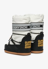 Bottes de neige noir et blanc avec une tige rembourrée, des lacets élastiques et une semelle en caoutchouc. Présente le logo brodé "SUPERDRY SNOW" en jaune.