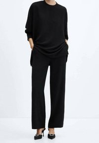 Top nero in maglia con scollo tondo e vestibilità ampia, abbinato a pantaloni neri a gamba larga. L'outfit presenta una texture liscia e un design minimalista.