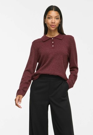 Femme aux cheveux raides et foncés porte un pull bordeaux à manches longues avec des boutons en perle et un pantalon large noir, debout devant un fond blanc.