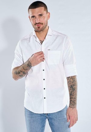 Man with tattoos wearing rolled-up white button-up shirt and blue jeans, adjusting shirt collar against plain light background.