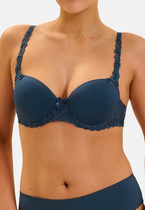 Femme portant un soutien-gorge rembourré bleu foncé avec des détails en dentelle sur les bretelles et la bande inférieure, et un sous-vêtement assorti sur un fond uni.
